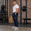 AI-generated original adult male powerlifter wearing J Navy W8WOLF Essentials Signature Joggers in an urban brick strength gym. Side logo, drawcord and cuff construction retained. White tee and gray shoes styling only. Other color supplier photos preserved.