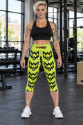 AI-generated original adult tattooed female bodybuilder wearing W8WOLF Electric Lime Oversized Wolf Yoga Capris in an urban strength gym. Mid calf cut, front waistband logo and original large black wolf heads with red eyes retained. Tank and shoes are styling only.