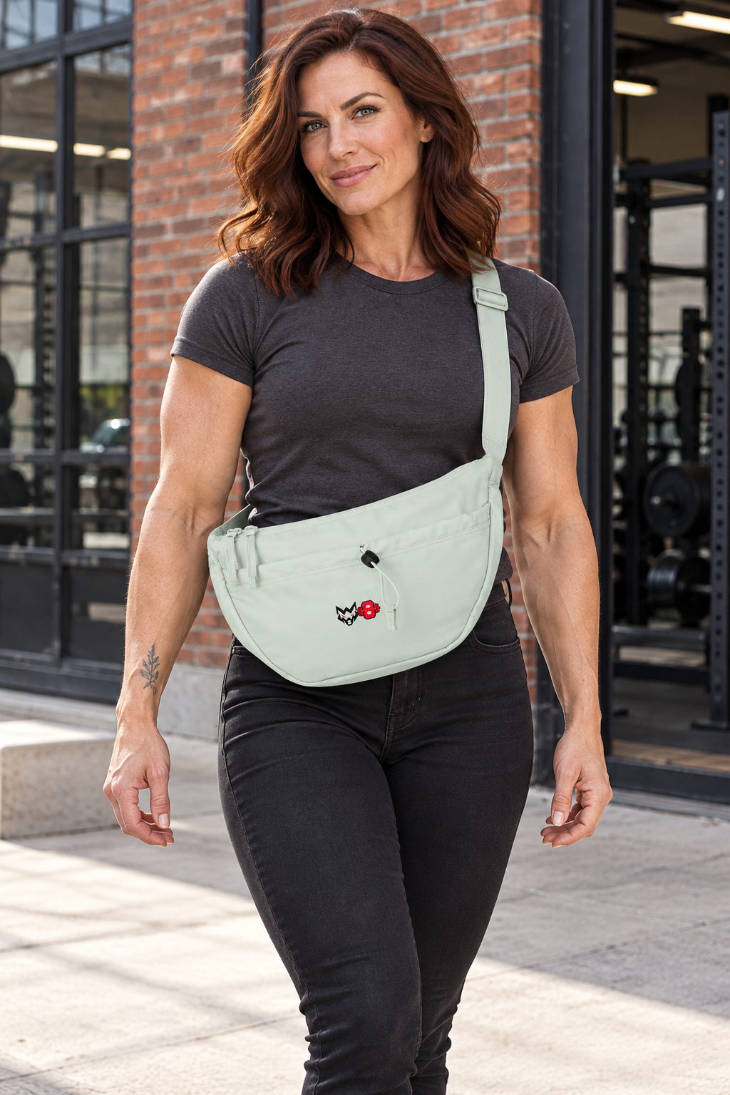 AI-generated original adult tattooed female bodybuilder wearing Fresh Green W8WOLF Emblem Crossbody Bag Pastels in an urban setting. Actual crescent construction front cord and compact original black wolf red eyes red barbell8 emblem retained. Clothing styling only; other bag colors preserved.