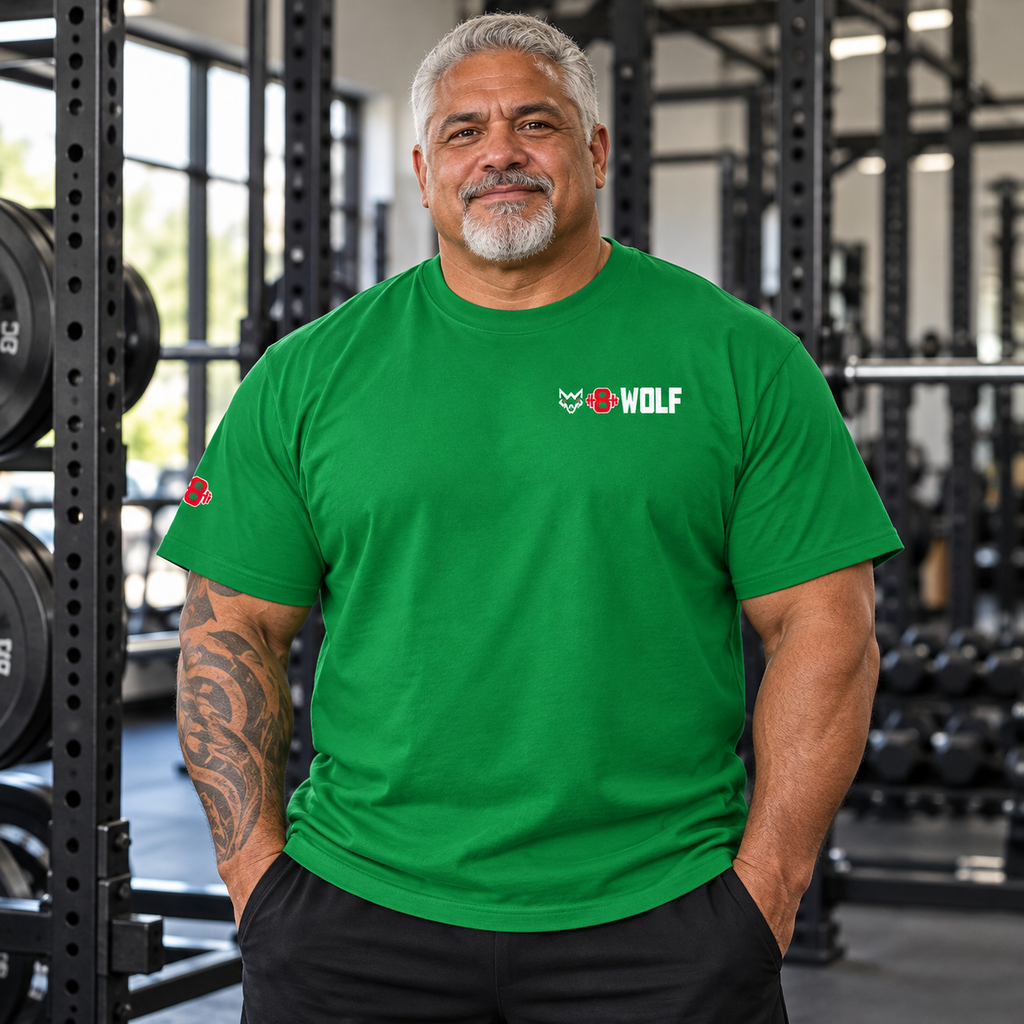 AI-generated original adult tattooed strongman wearing W8WOLF Max Heavyweight Tee Kelly Green in an urban gym. Actual green cotton cut small white wolf red eyes red barbell8 WOLF left chest and right sleeve red8 retained. Pants styling only.