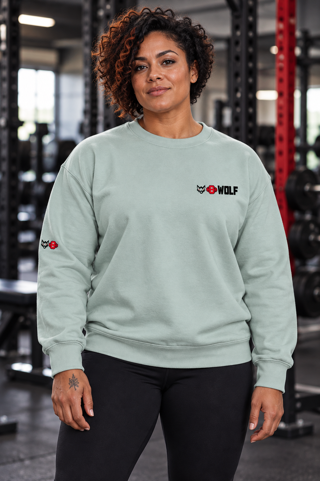AI-generated original adult female powerlifter wearing W8WOLF Signature Crewneck Agave in an urban gym. Actual pale sage sweatshirt cut small black wolf red eyes red barbell8 WOLF left chest and right sleeve wolf8 retained. Pants styling only.